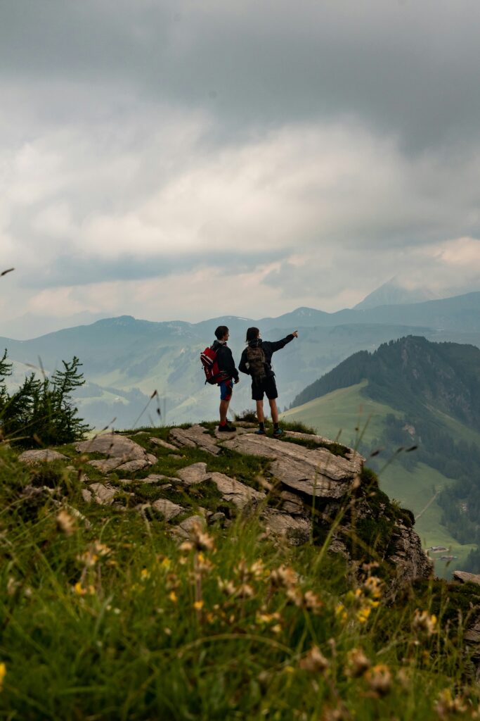 Two people standing atop a mountain in the daytime, looking out over mountains in the distance. One person is pointing ahead toward the horizon while the other stands with their hands at their sides.